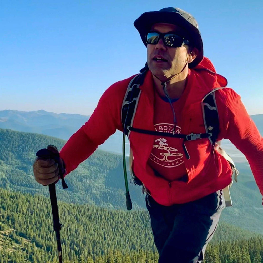 Hiker wearing red Kyoto Botanicals logo t-shirt and jacket on a mountain trail with forested Colorado peaks in the background — Pure Harmony outdoor lifestyle.