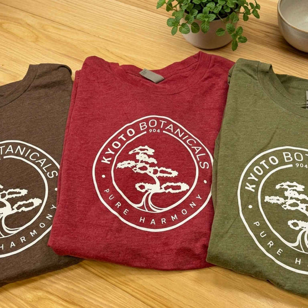 Kyoto Botanicals limited-edition folded T-shirt trio on natural wood—Espresso, Cardinal Red, and Military Green nature-inspired wellness apparel.